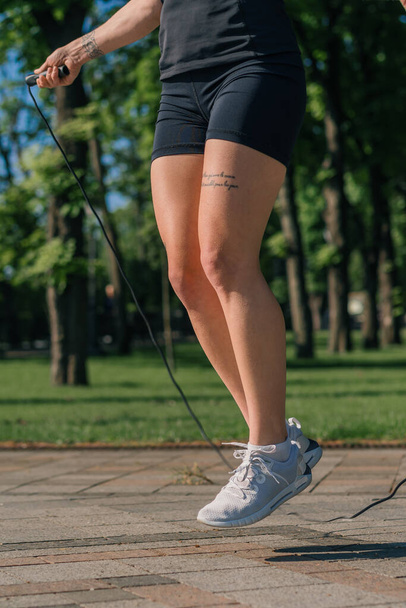 Fitness young sportswoman girl in the morning doing sports jumping rope during exercise outdoors in the city in health park close-up of legs - 写真・画像