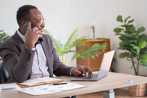Egy fekete öltönyös afro-amerikai üzletember mobiltelefont használ, hogy kommunikáljon az ügyfelekkel és egy laptopot, hogy megtekinthesse a cég profitját. bizalomépítés az ügyfelek számára az irodába való befektetés érdekében. - Fotó, kép