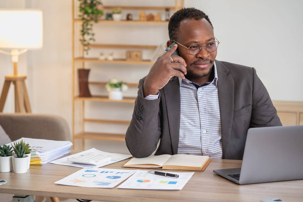 Afrikkalainen amerikkalainen liikemies mustassa puvussa käyttää matkapuhelinta kommunikoidakseen asiakkaiden kanssa ja kannettavaa tietokonetta nähdäkseen yrityksen voitot. rakentaa luottamusta asiakkaiden tulla ja investoida toimistoon. - Valokuva, kuva