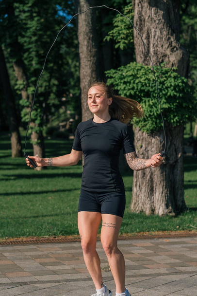 Fitness young sportsman girl with tattoos in the morning doing sports jumping rope during exercise outdoors in the city in health park - Photo, Image