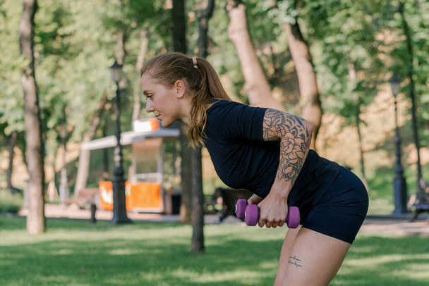 портрет молодої задоволеної спортивної дівчини, яка виконує вправи з гантелями на відкритому повітрі під час тренування в парку концепція здорового способу життя - Фото, зображення
