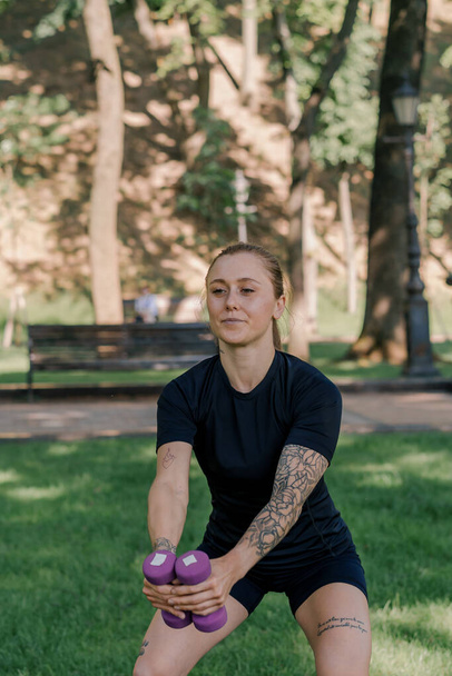 портрет молодой довольной спортивной девушки, делающей упражнения с гантелями на открытом воздухе во время тренировки в парке концепции здорового образа жизни - Фото, изображение