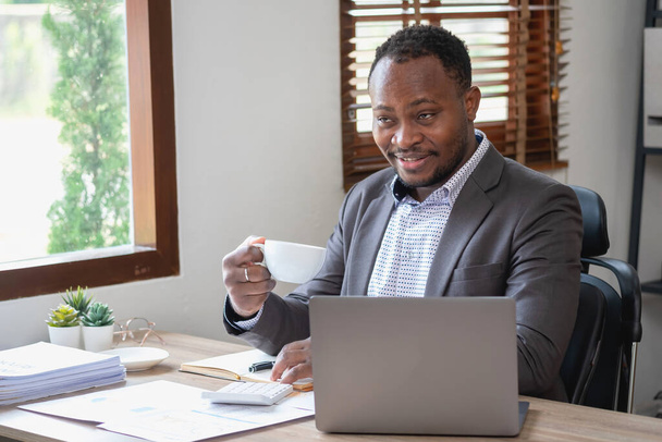 Afroamerikanischer Geschäftsmann im schwarzen Anzug hält eine Tasse Kaffee in der Hand, um sich zu entspannen, nachdem er die Arbeit des Unternehmens auf einem Laptop im Büro überprüft hat. - Foto, Bild