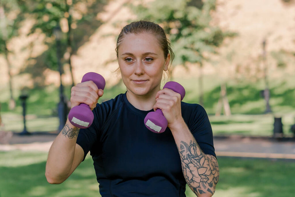 portrait of a young satisfied sports girl doing exercises with dumbbells outdoors during a workout in park healthy lifestyle concept - Photo, Image