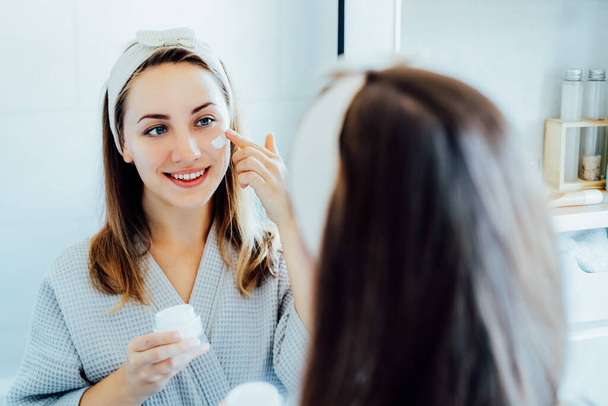 Young woman in bathrobe looking in the mirror, holding bottle with moisturizer cream and applying it on face. Skin hydrating. Cosmetic spa procedures. Beauty self-care at home. Selective focus - Photo, image