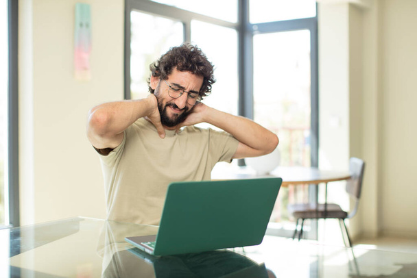 jovem homem barbudo adulto com um laptop sentindo-se ansioso, doente, doente e infeliz, sofrendo uma dor dolorosa no pescoço - Foto, Imagem