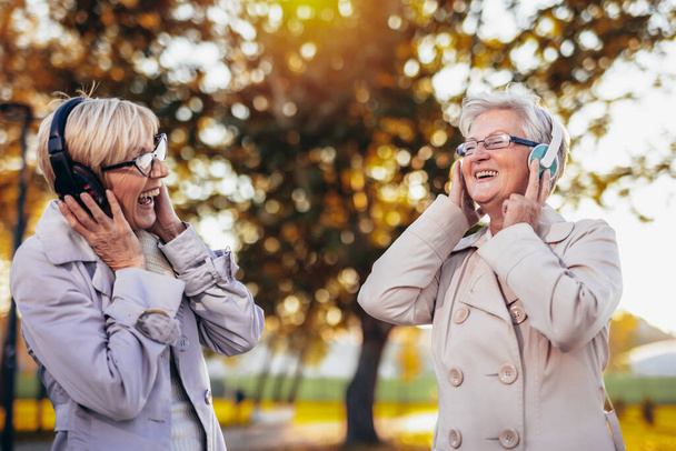 秋のカラフルな公園で彼らのワイヤレスヘッドフォンで音楽を聴く2人の年上の女性 - 写真・画像