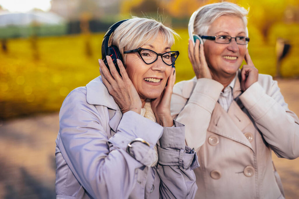 Δύο μεγαλύτερες γυναίκες ακούγοντας μουσική στα ασύρματα ακουστικά τους στο πολύχρωμο πάρκο το φθινόπωρο - Φωτογραφία, εικόνα