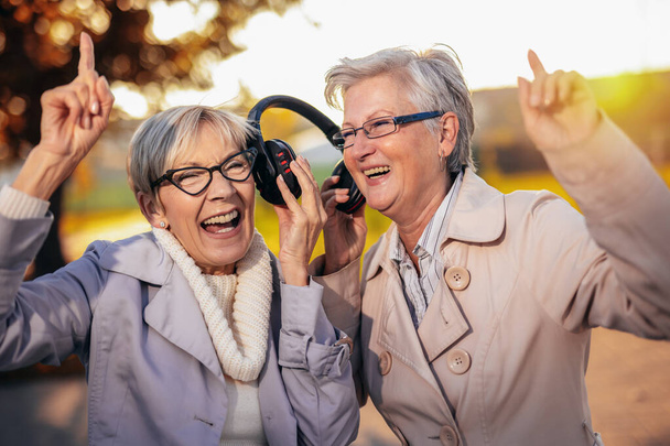Две пожилые женщины слушают музыку на беспроводных наушниках в красочном парке осенью - Фото, изображение