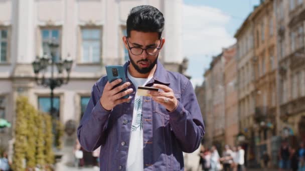 Iloinen nuori intialainen mies, joka käyttää luottokortin älypuhelinta siirtäessään rahaa, tehdessään ostoksia verkossa, tilatessaan ruokaa, varatessaan hotellihuoneen. Hindu kaveri kävelee kaupunkien kadulla ulkona - Materiaali, video