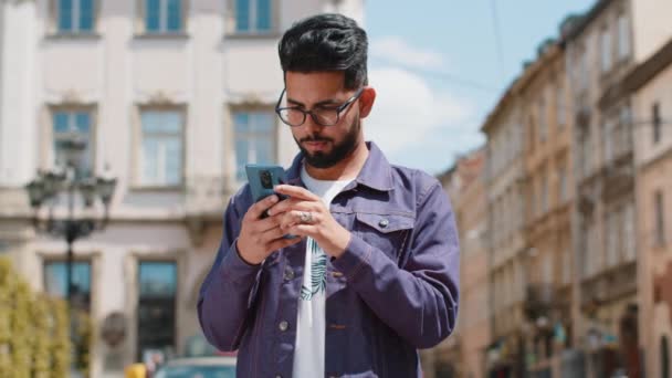 Lachende jonge indiaanse man met behulp van smartphone typen sms-berichten surfen op internet, afwerking van het werk, kijken naar de camera. Baard hindoe man lopen in stedelijke stad straat achtergrond. Stadslevensstijl buiten - Video