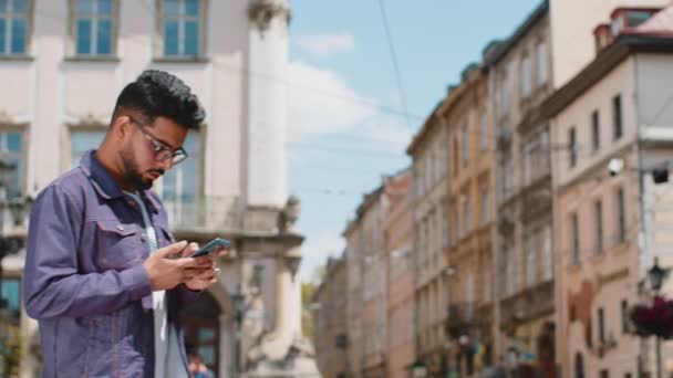 Tourist traveler young adult indian man using smartphone typing text messages looking for search a way on map in mobile navigator app outdoors. Hindu guy walking passes by urban city street background - Footage, Video