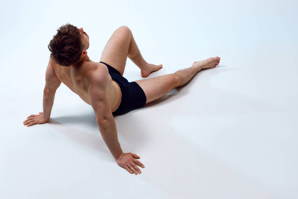 Young man with athletic, muscular, fit body lying on floor, posing shirtless in underwear against white studio background. Concept of mans beauty, sportive and healthy lifestyle, athletic body - Foto, afbeelding