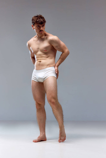 Full-length image of young man with healthy, strong, muscular body posing shirtless in underwear against grey studio background. Concept of mans beauty, sport, health, athletic body, medicine - Photo, Image