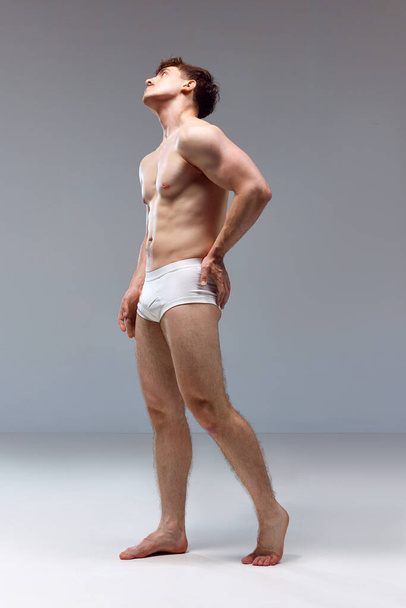 Full-length image of young man with healthy, strong, muscular body posing shirtless in underwear on grey studio background. Back pains. Concept of mans beauty, sport, health, athletic body, medicine - 写真・画像