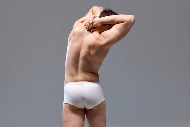 Rear view of muscular, relief male back. Man posing shirtless in underwear against grey studio background. Stretching. Concept of mans beauty, sport, health, athletic body, medicine - Photo, image