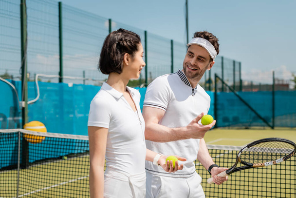 positive man and woman holding tennis balls and racket on court, hobby and leisure - Photo, Image