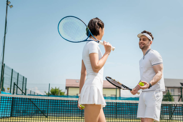 positive couple holding tennis balls and rackets on court, hobby and summer leisure, sport - Zdjęcie, obraz