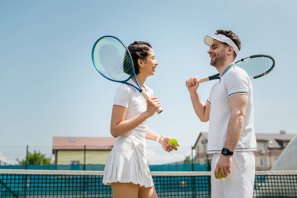 feliz hombre y mujer en ropa deportiva charlando mientras está de pie con raquetas de tenis y pelotas en la cancha - Foto, Imagen