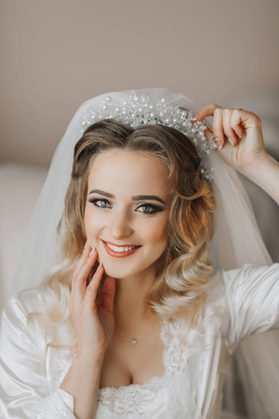 A blonde bride is wearing a satin robe and sitting in her room, posing and showing off her tiara and veil. Beautiful hair and make-up, open bust. Wedding portrait. Sincere smile - Photo, image