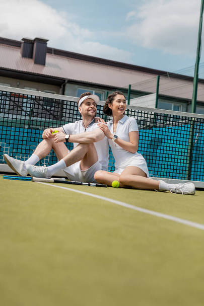veselý muž a žena sedí v blízkosti tenisové sítě, rakety a míč, letní sport, volný čas a zábava - Fotografie, Obrázek