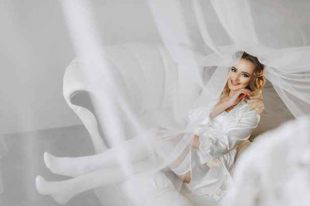 Blonde bride wearing a satin robe and lying on a couch, posing and showing off her tiara and veil. Beautiful hair and make-up, open bust. Wedding portrait. Sincere smile - 写真・画像