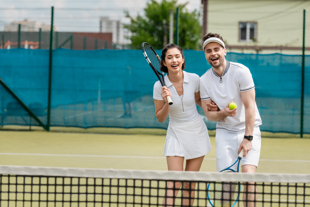 cheerful sporty couple in active wear holding rackets and ball near tennis net, hobby and sport - Foto, Imagem