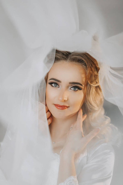 The blonde bride is dressed in a satin robe and lies on a sofa, posing under a veil. Beautiful hair and make-up, open bust. Wedding portrait. Sincere smile - Foto, Bild