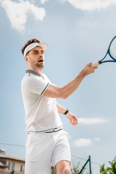 handsome man in visor cap and active wear holding racket on tennis court, backhand, hobby and sport - Foto, imagen