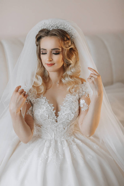 the bride in a white wedding dress. happy beautiful young woman in white traditional wedding dress in her room on leather sofa holding veil - Foto, Bild