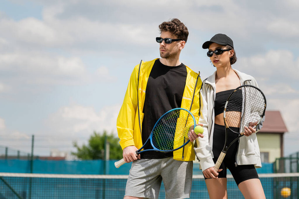 Sommersport, Mann und Frau mit Sonnenbrille stehen mit Schlägern auf dem Tennisplatz, Sportlermode - Foto, Bild
