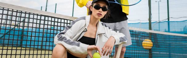 Pankart, güneş gözlüklü ve şapkalı bir kadın, adamın yanında poz veriyor, top ve tenis raketi tutuyor, etkin giyiniyor. - Fotoğraf, Görsel