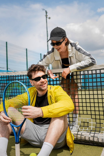 χαρούμενη γυναίκα με γυαλιά ηλίου και ενεργητική ένδυση μιλώντας σε άνδρα με ρακέτα τένις, δίχτυ τένις, αθλητισμό - Φωτογραφία, εικόνα