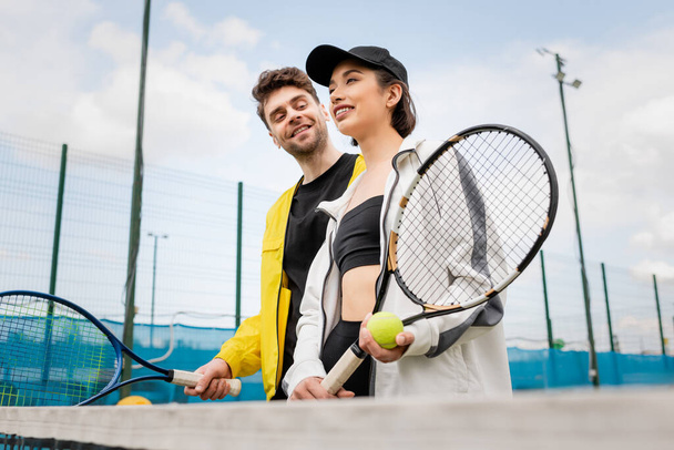 couple joyeux en tenue active élégante tenant raquettes de tennis et ballon sur le court, mode de vie et sport - Photo, image