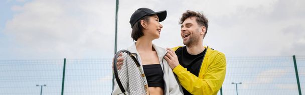 baner, pozytywny mężczyzna w aktywnym nosić przytulanie kobieta w czapce z rakietą tenisową na korcie, styl życia - Zdjęcie, obraz