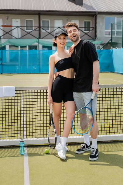 romantika na tenisovém kurtu, pozitivní pár v černém aktivní opotřebení stojící s tenisovými raketami u míče - Fotografie, Obrázek