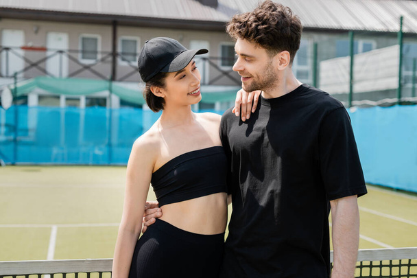 pozitivní pár v černém aktivní oblečení stojící v blízkosti tenisové sítě, romantika na tenisovém kurtu - Fotografie, Obrázek