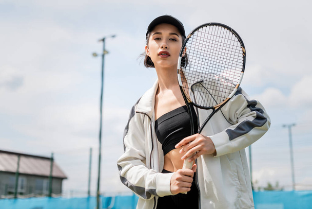 sporty woman in active wear and cap looking at camera and holding tennis racket on court, sport - Zdjęcie, obraz