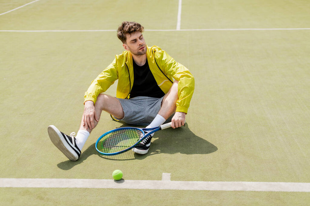 przystojny mężczyzna w aktywnym ubraniu spoczywa na korcie tenisowym, męski tenisista z rakietą, sport - Zdjęcie, obraz