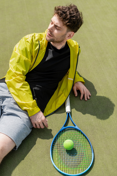 tiro en la cabeza, hombre guapo descansando cerca de pelota de tenis y raqueta, jugador de tenis masculino en la cancha - Foto, Imagen