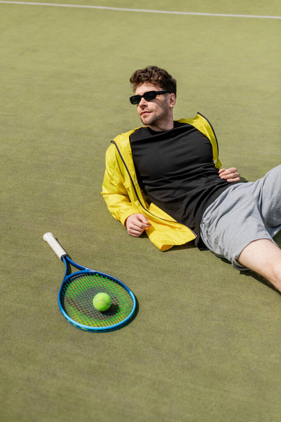 красивий чоловік в сонцезахисних окулярах відпочиває біля тенісного м'яча і ракетки, чоловічий тенісист, спорт і стиль - Фото, зображення