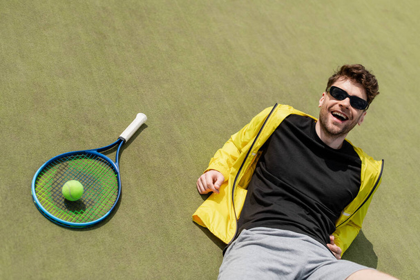 ευτυχισμένος άνθρωπος με γυαλιά ηλίου αναπαύεται κοντά σε μπάλα του τένις και ρακέτα, αρσενικό παίκτη του τένις, τον αθλητισμό και το στυλ - Φωτογραφία, εικόνα