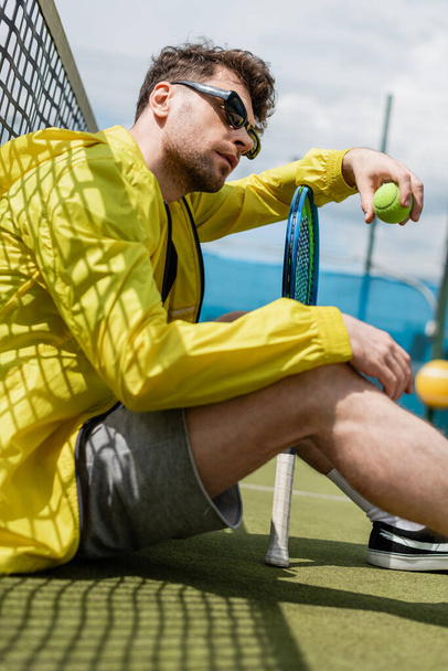 jugador masculino en gafas de sol sentado cerca de la red de tenis, celebración de pelota y raqueta de tenis, deporte y estilo - Foto, Imagen