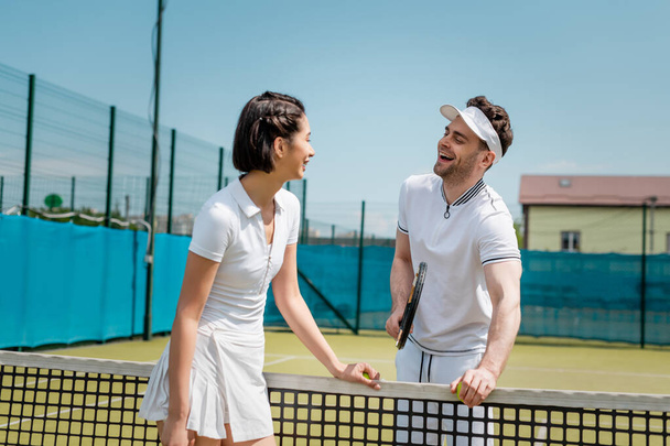 homme heureux regardant une femme près d'un filet de tennis, couple joyeux debout sur un court de tennis, vêtements actifs - Photo, image