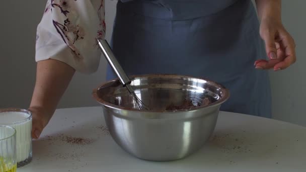 Una mujer agrega un vaso de leche a la masa para una magdalena, pastel o brownie en un lazo de metal. La preparación de la masa para el pastel de cumpleaños. Paso para hacer magdalenas, tarta, bizcocho. Proceso de cocción paso a paso - Imágenes, Vídeo