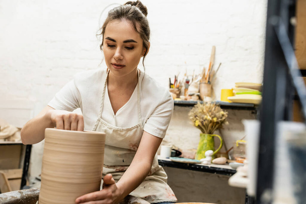 Brunette craftswoman in apron making clay vase and working on pottery wheel in ceramic workshop - Photo, Image