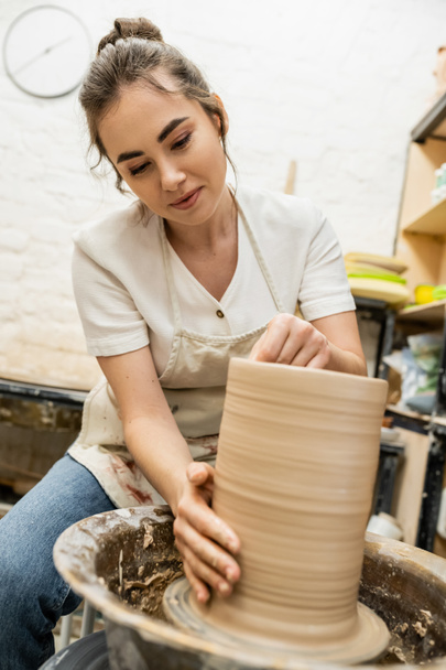 Brunette female potter in apron making clay vase on pottery wheel in workshop at background - Photo, Image