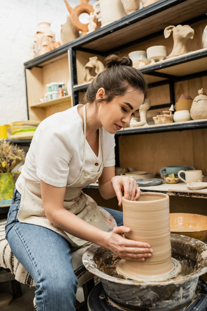 バックグラウンドでワークショップで陶器ホイールに粘土彫刻を作成するエプロンのブルネット女性ポッター - 写真・画像