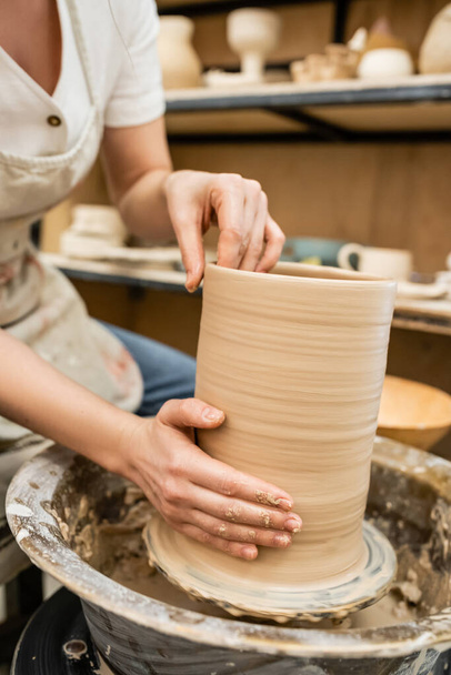 Cropped view of craftswoman in blurred apron making clay vase on pottery wheel in ceramic studio - Photo, Image