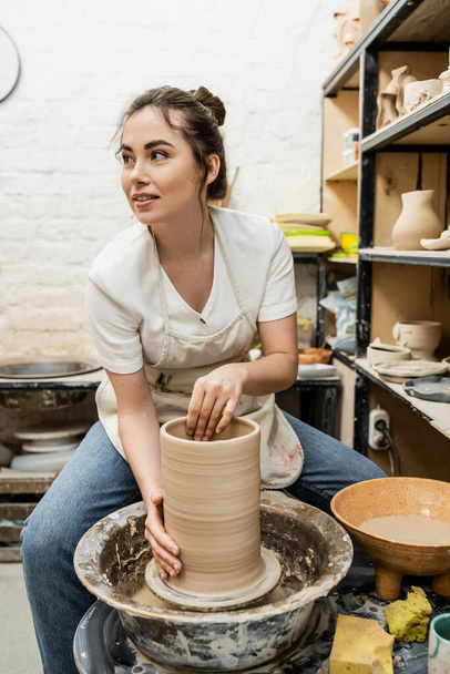 Artesana morena sonriente en delantal haciendo jarrón de barro sobre rueda de cerámica en taller - Foto, Imagen